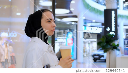 Young muslim businesswoman in hijab enjoying coffee break, working on tablet while walking through modern shopping mall interior 129440541