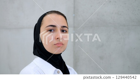 Professional muslim female healthcare worker wearing hijab and white coat, standing against concrete wall, displaying confident facial expressions while facing camera 129440542