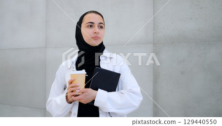 Young muslim woman wearing hijab and white shirt, holding takeaway coffee cup and notebook, standing contemplatively against modern concrete wall background Young muslim woman wearing hijab and white shirt, holding takeaway coffee cup and notebook, standing contemplatively against modern concrete wall background 129440550