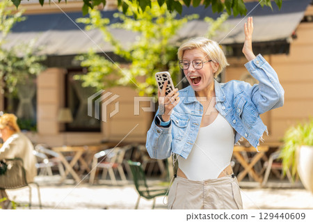 Excited European woman using mobile phone typing browsing celebrating win on urban city street Excited European woman using mobile phone typing browsing celebrating win on urban city street 129440609