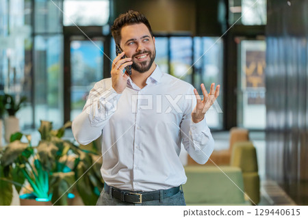 Smiling Caucasian young businessman talking on smartphone discussing work in office hotel lobby 129440615