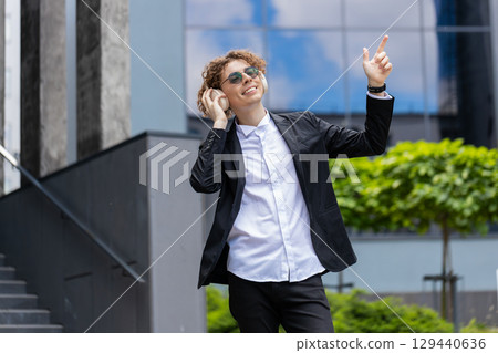 Happy relaxed young Caucasian businessman in wireless headphones dancing, listening music outdoors 129440636