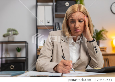 Upset mature businesswoman using calculator feeling worried about financial problem at office desk 129440655