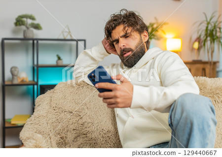 Upset displeased Indian man guy using smartphone reading bad news sitting on couch in living room 129440667