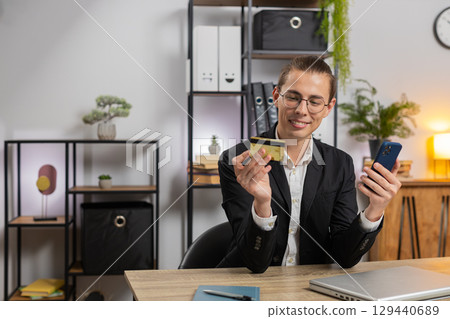Happy young businessman using bank credit card and smartphone for online shopping at home office 129440689