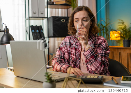 Businesswoman girl sitting at workplace desk drinking water while working with laptop in home office 129440715