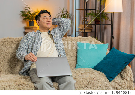 Happy mature Chinese man sitting on couch closing laptop after finishing work in living room at home 129440813