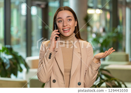 Smiling Caucasian young businesswoman talking on smartphone discussing work in office hotel lobby Smiling Caucasian young businesswoman talking on smartphone discussing work in office hotel lobby 129441030