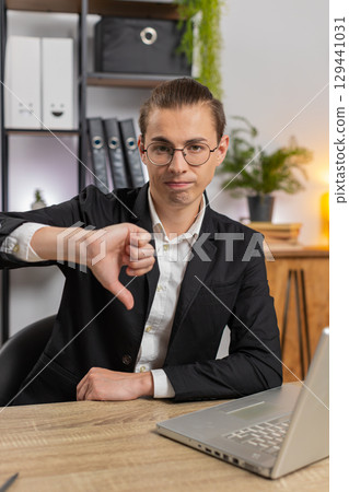 Upset young Caucasian businessman freelancer showing double thumbs down sitting at home office desk 129441031