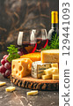 A wooden tray with various sliced cheeses against a backdrop of two glasses and a bottle of red wine in a dark interior. 129441093
