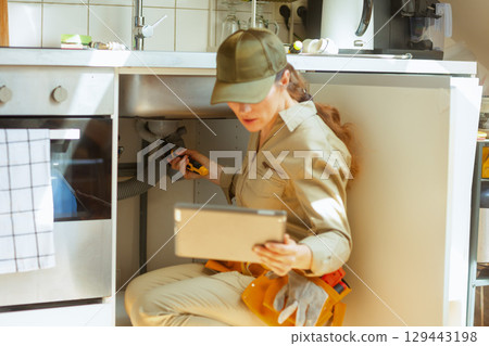 Plumber inspects pipe under sink using tablet and wrench 129443198