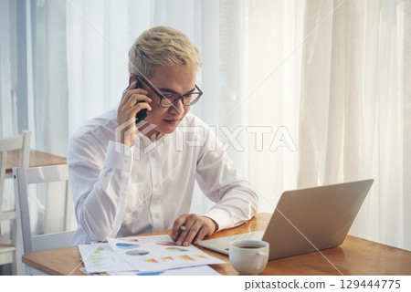 Asian Businessman using laptop calling phone at office desk. Man wear eye glasses talking smartphone read financial graph chart Planning marketing info. Happy Asian business people work office firm 129444775