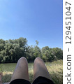 Relaxing Summer Day by the River. First Person View of Legs Resting on Grass, Peaceful Nature, Blue Sky, Green Water and Trees, Outdoor Vacation Scene, Calm and Freedom Mood Concept 129451047