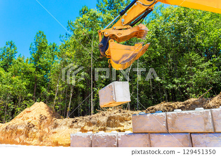 Heavy machinery is lifting concrete block at construction site surrounded by trees under build retaining walls Heavy machinery is lifting concrete block at construction site surrounded by trees under build retaining walls 129451350