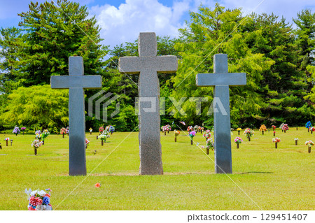 Three upright stone crosses are placed in tranquil cemetery with colorful flower arrangements under bright sky. Three upright stone crosses are placed in tranquil cemetery with colorful flower arrangements under bright sky. 129451407