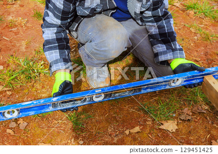 Worker checks leveling of wooden structure while kneeling on grass, ensuring precision in building process. Worker checks leveling of wooden structure while kneeling on grass, ensuring precision in building process. 129451425