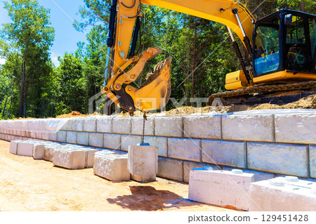 Excavator positioned next to wall lifts concrete blocks during construction in forested area under build retaining walls 129451428