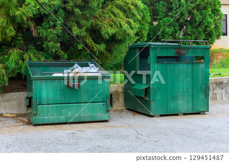 Two green dumpsters are located next to concrete wall, filled with recyclable materials, under brighter sunlight. 129451487