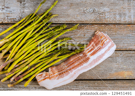 Fresh green asparagus spears sit beside raw bacon on weathered wooden surface, ready for cooking preparation. 129451491