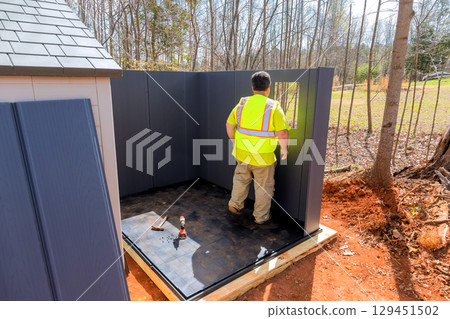 Construction worker in bright vest in forested area under assembling storage shed 129451502