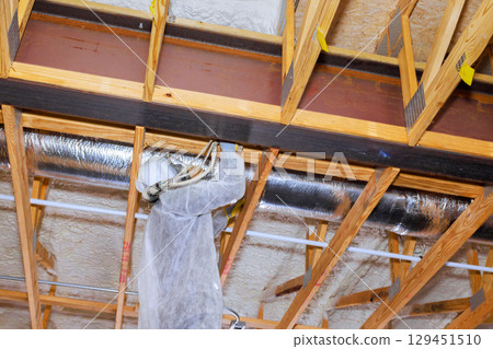 Construction worker skillfully installs spray foam insulation in home renovation on wooden beams structure 129451510