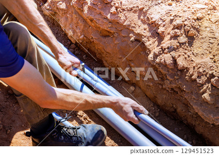 Construction worker carefully positions plumbing pipes into trench on works day. Construction worker carefully positions plumbing pipes into trench on works day. 129451523