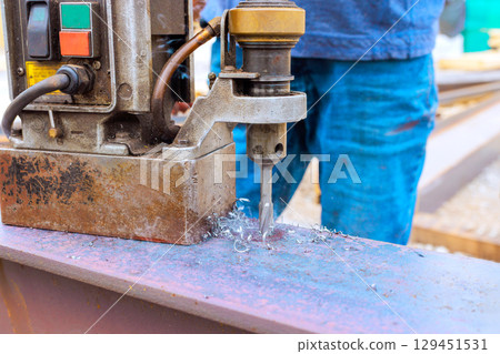 Worker operates metal drilling machine on steel beams while shavings fly everywhere in an industrial area. 129451531