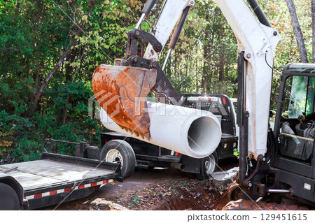 Construction workers use an excavator to lift concrete drainage pipe from truck for utility installation in forest. 129451615