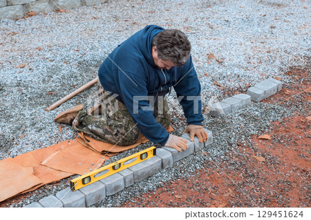 Worker carefully lays bricks for pathway, ensuring level placement on gravel surface during fall season. Worker carefully lays bricks for pathway, ensuring level placement on gravel surface during fall season. 129451624