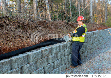 Worker arranges stone blocks for retaining wall by gravel road amid trees. 129451630
