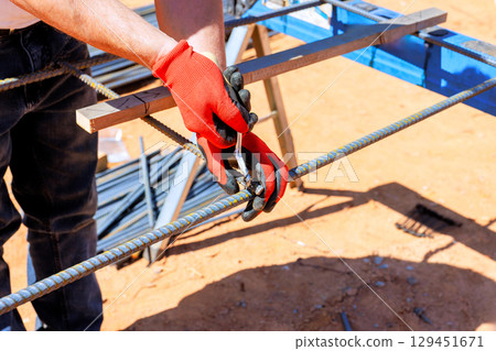 Worker in gloves grips metal rebar with pliers while constructing steel rebar framework 129451671