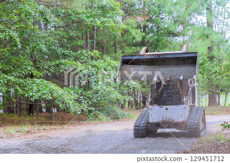 Skid uploader distributes gravel along dirt path in shaded forest, enhancing access for leveling landscaping 129451712