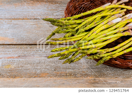 Freshly harvested green asparagus spears rest in woven basket on weathered wooden surface. 129451764