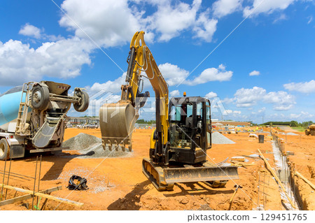 Excavator lifts dirt while concrete mixer pours materials at construction site under works day 129451765
