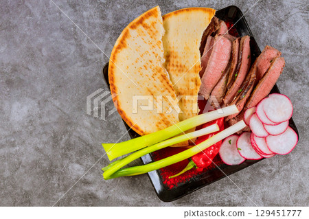 Slices of tender beef alongside flatbread, radishes, green onions arranged artfully on black platter. Slices of tender beef alongside flatbread, radishes, green onions arranged artfully on black platter. 129451777