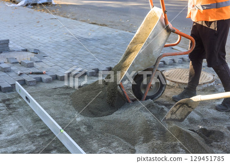 Construction worker pours sand from wheelbarrow onto ground to prepare for paving bricks during works day. Construction worker pours sand from wheelbarrow onto ground to prepare for paving bricks during works day. 129451785