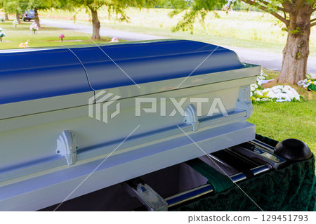 White casket is positioned in cemetery during funeral service among green trees floral tributes. 129451793