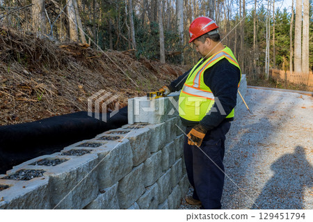 Construction worker measures stone blocks while working on retaining wall beside gravel road 129451794