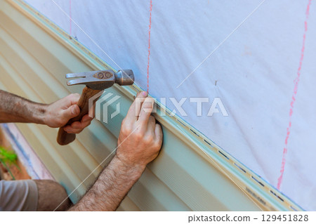Contractor is using hammer to secure facade vinyl siding plank on exterior wall of house under renovation. 129451828