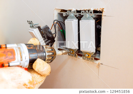 An electrician adjusts wall switches using power drill while ensuring proper installation safety. 129451856