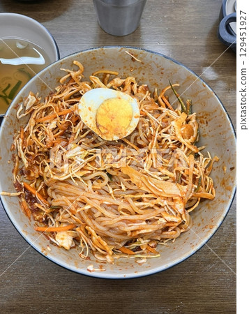 Bibimyeon, a flavorful Korean dish featuring cold noodles tossed in a spicy, tangy sauce, topped with fresh vegetables and a boiled egg. Perfect for summer meals, showcasing vibrant flavors and colors Bibimyeon, a flavorful Korean dish featuring cold noodles tossed in a spicy, tangy sauce, topped with fresh vegetables and a boiled egg. Perfect for summer meals, showcasing vibrant flavors and colors 129451927