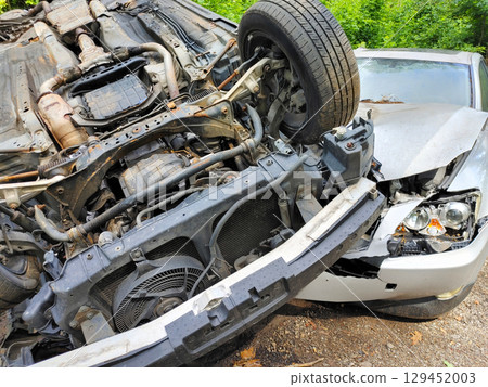 Flipped car is leaning against another vehicle in wooded area following collision, extensive damage. 129452003