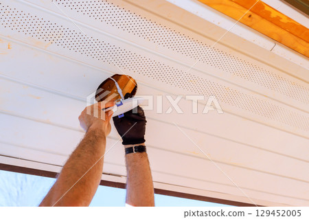 Homeowner applies lighting fixture in ceiling in soffit under eaves of house in suburb. Homeowner applies lighting fixture in ceiling in soffit under eaves of house in suburb. 129452005