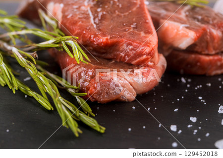 Raw meat steaks with fresh herbs, coarse salt are showcased on dark slate surface, ready preparation. Raw meat steaks with fresh herbs, coarse salt are showcased on dark slate surface, ready preparation. 129452018