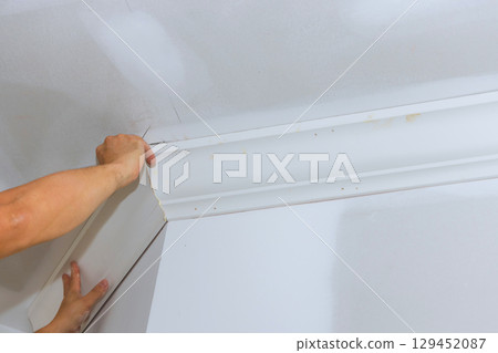 Carpenter carefully aligns, installs crown molding at corner of white room to enhance its appearance design. 129452087