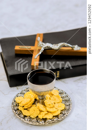 Silver cup filled with wine plate of wafers placed by Bible and crucifix during morning prayer. 129452092