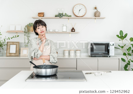一位繫著圍裙的年輕亞洲女子在廚房的電磁爐前做飯，思考著，憂心忡忡 129452962
