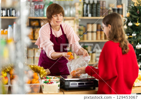 Woman sells fruits to teen girl, puts tangerines in bag, puts them on scale and weighs fruits Woman sells fruits to teen girl, puts tangerines in bag, puts them on scale and weighs fruits 129453067