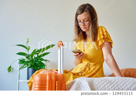 Woman in dress sitting on bed with orange suitcase using smartphone indoors Woman in dress sitting on bed with orange suitcase using smartphone indoors 129453115