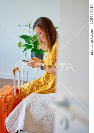 Woman sitting on bed with suitcase and using smartphone Woman sitting on bed with suitcase and using smartphone 129453116